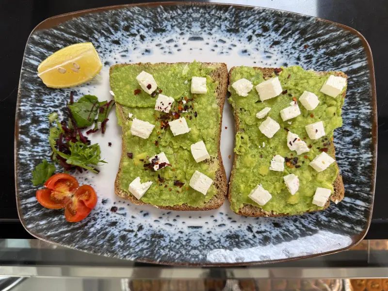 Smashed avocado and crumbled feta over two slices of toasted sourdough, scattered with chilli flakes, with lemon, salad leaves and tomato.
