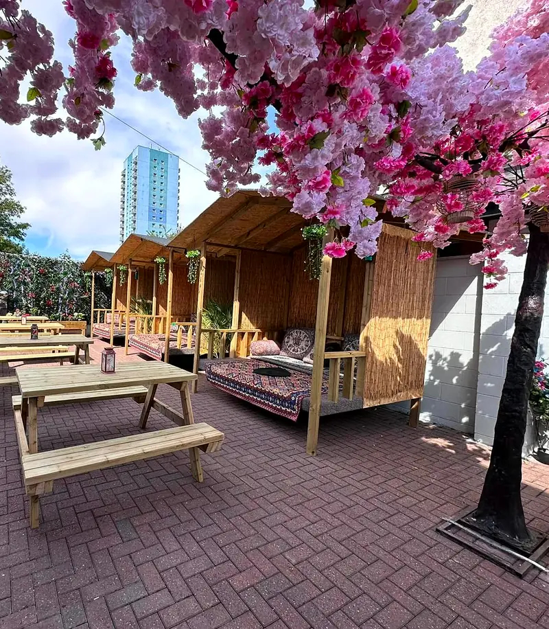 Rumi's back garden: wooden takht cabanas with Persian rugs and picnic benches, under a canopy of pink blossom.